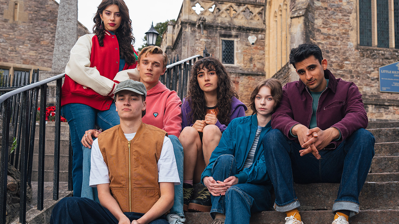 "Six young people sit and pose on historic stone steps outside an old church building, dressed in colorful casual outfits, creating a moody and thoughtful atmosphere in a picturesque outdoor setting."